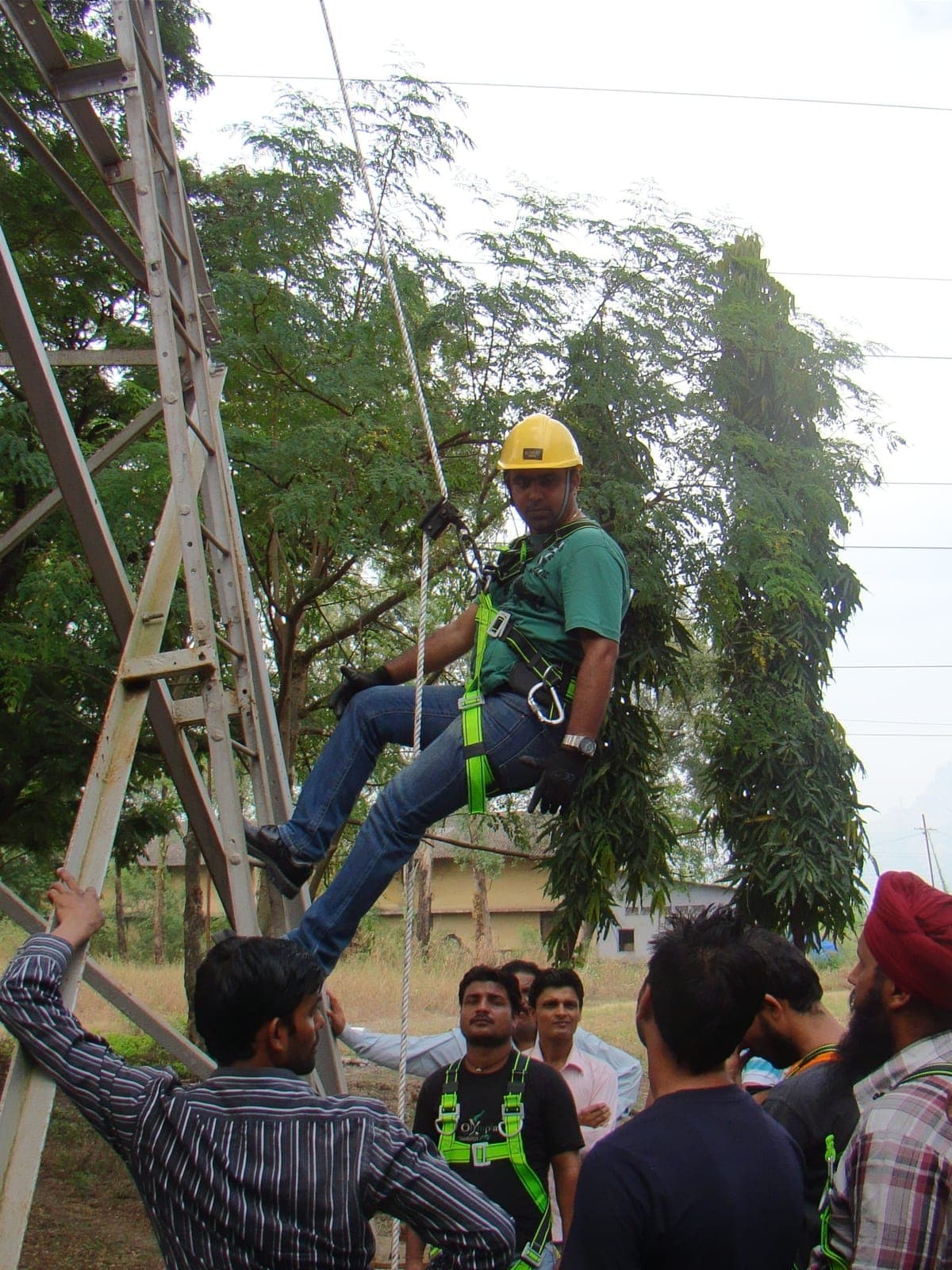 Trainee in a yellow hard hat and full-body harness suspended on a rope beside a steel training tower at the Eduforce campus, with fellow trainees watching from below