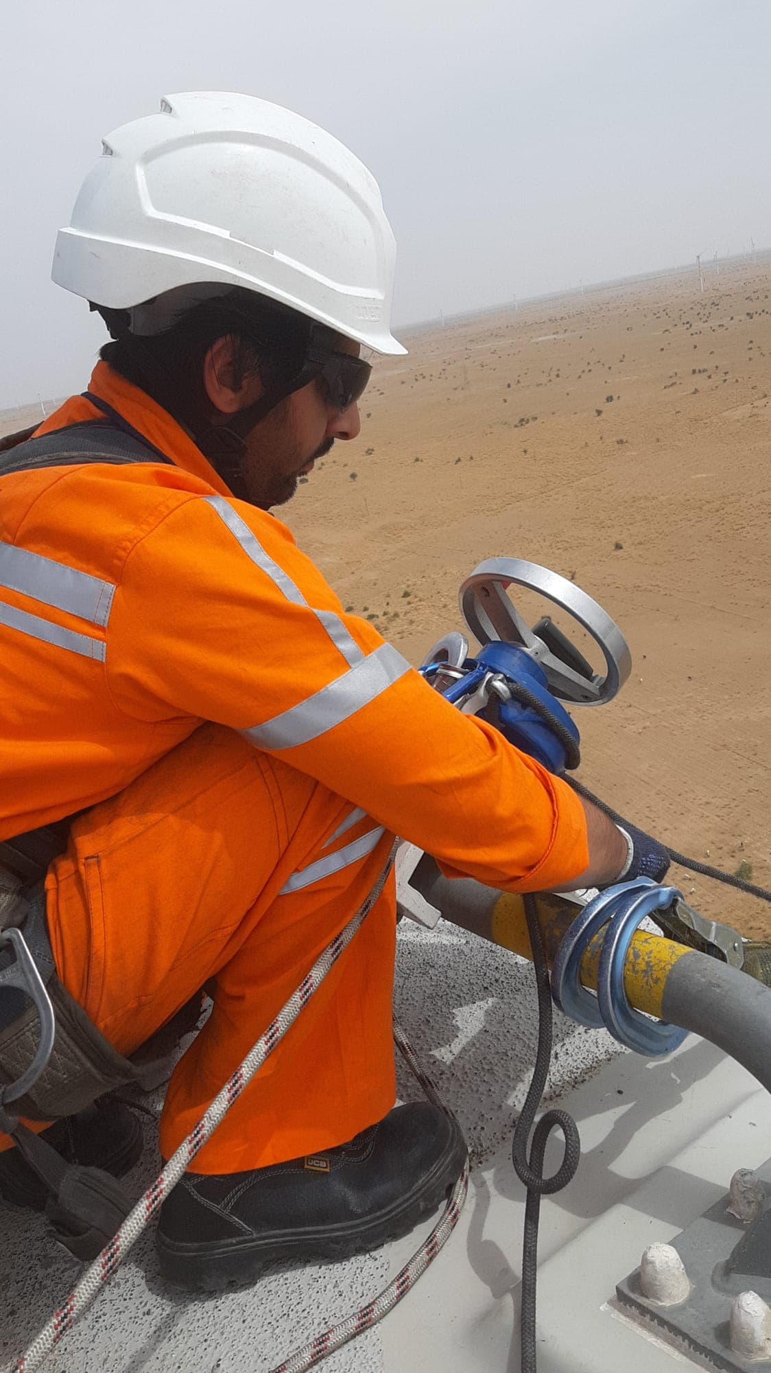 Technician in orange high-visibility coveralls rigging a rope through an anchor point on a wind turbine nacelle roof