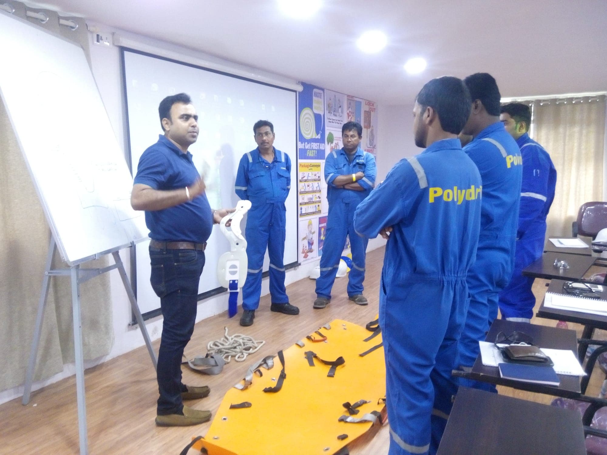 Trainer briefing participants in blue coveralls beside a basket stretcher laid on the floor