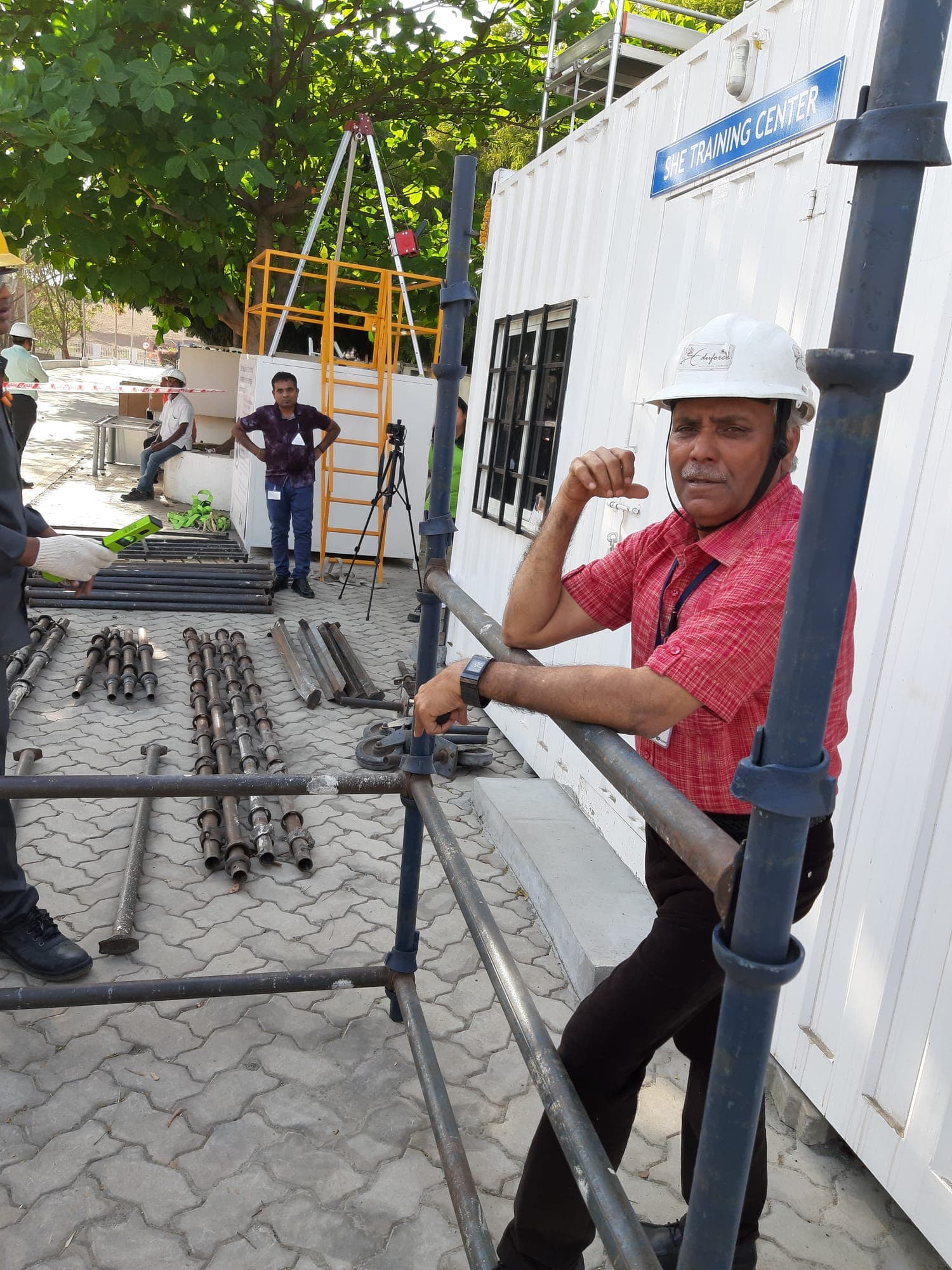 Trainer in a hard hat explaining a coupler connection on a part-built scaffold bay, with scaffold tubes and fittings laid out on the ground
