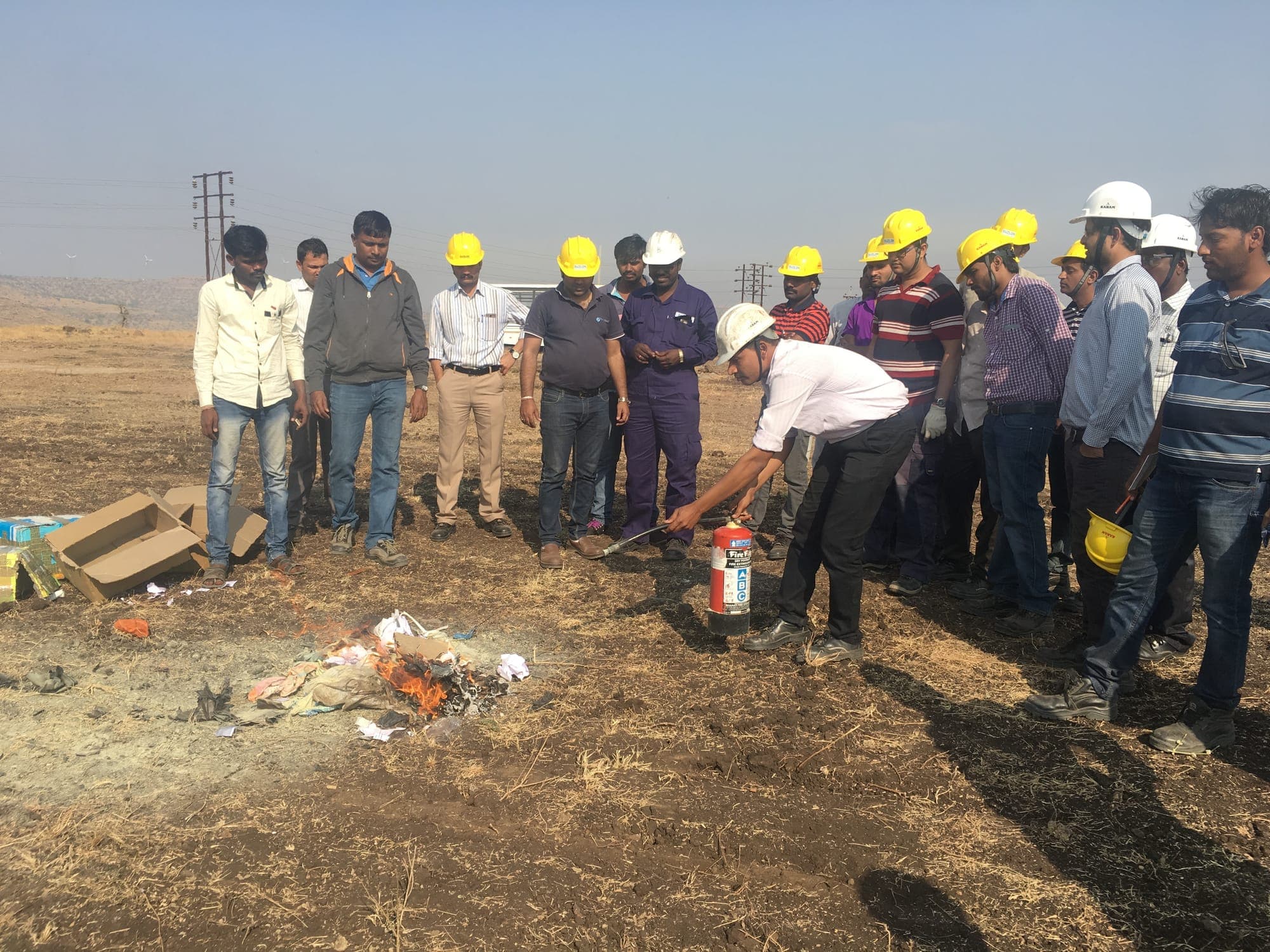 Trainee discharging a dry powder extinguisher on open ground while a line of colleagues in hard hats observes