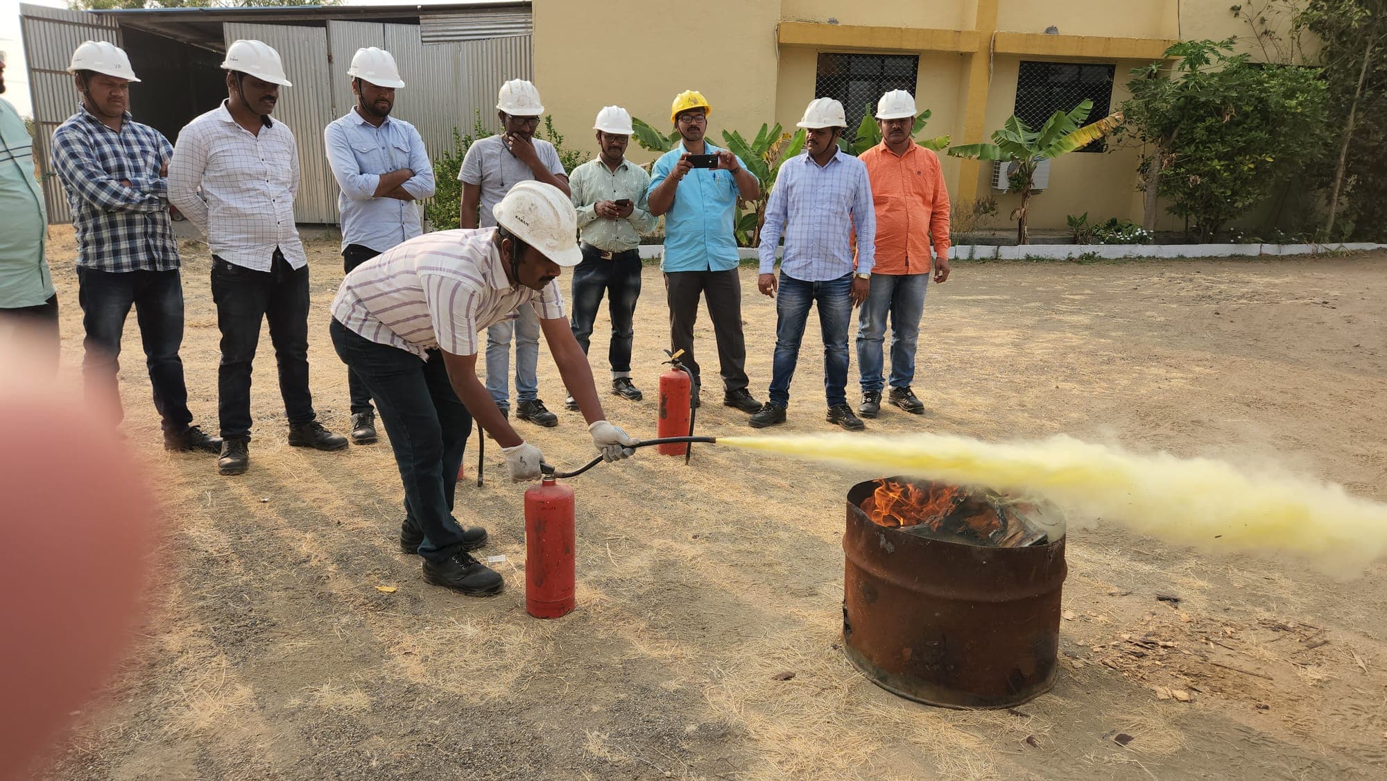 Trainee discharging an extinguisher at a drum fire under supervision while colleagues in hard hats observe
