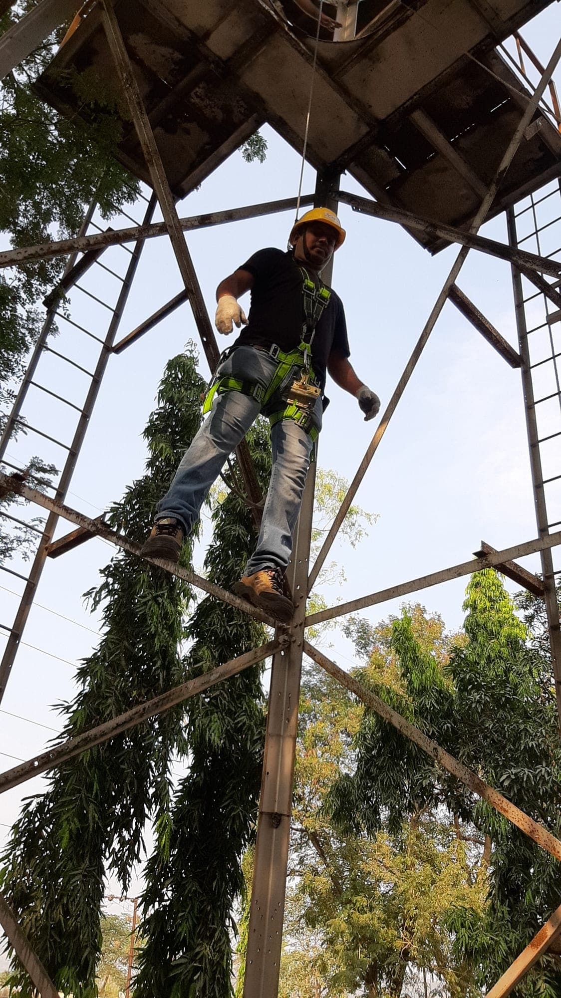 Trainee in a helmet, full-body harness and shock-absorbing lanyard standing on a cross-member of a steel lattice tower