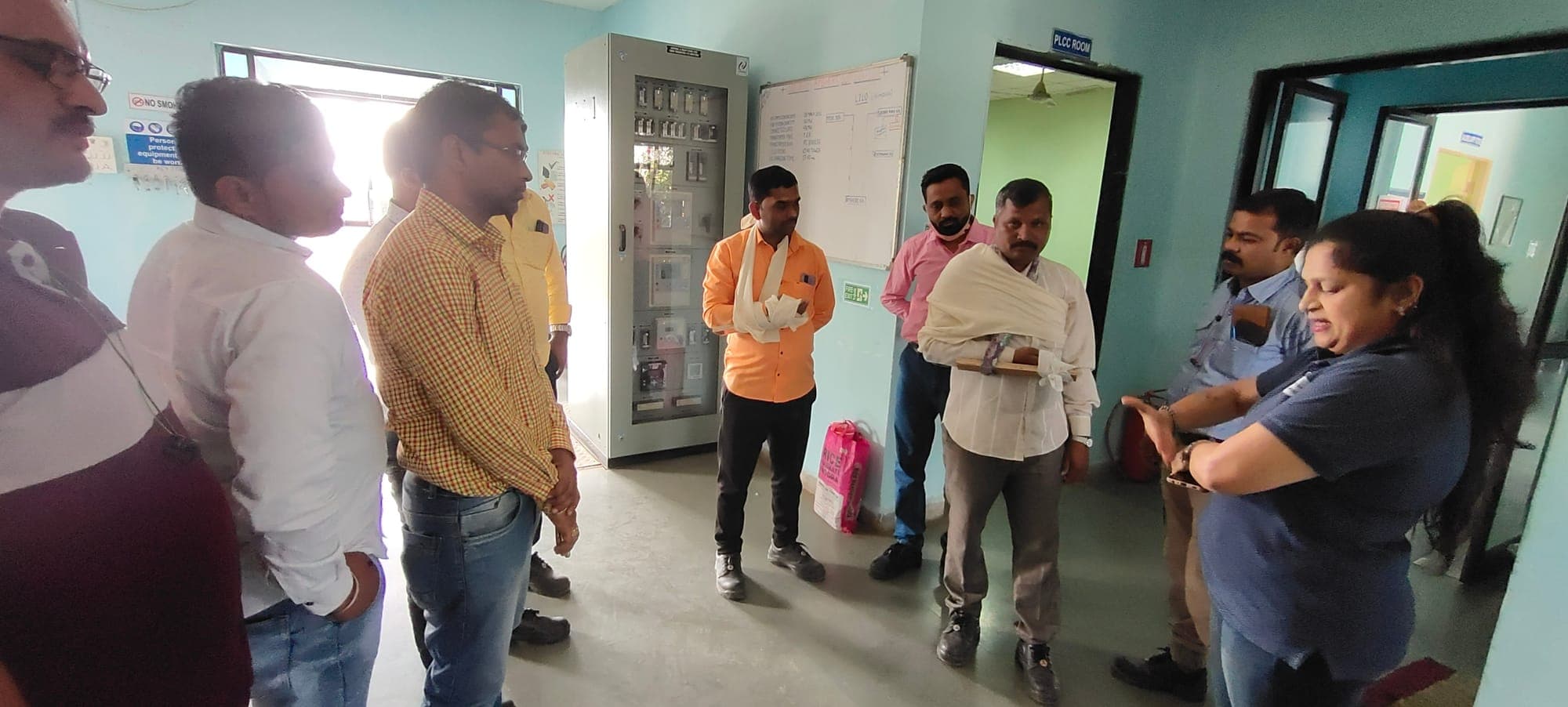 Trainer addressing a group in a plant corridor, with two participants wearing arm slings and a splint fitted during the exercise