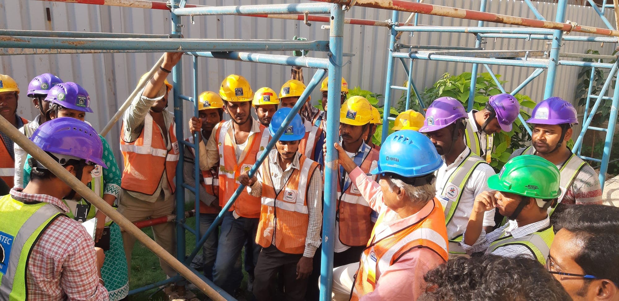 Large group of construction workers in coloured helmets gathered around a scaffold for a safety talk