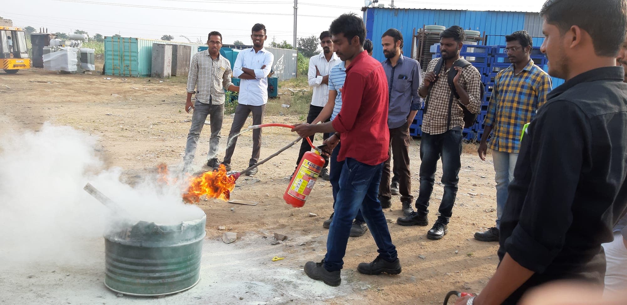 Trainee discharging a dry powder extinguisher into a burning drum while colleagues observe during live fire training