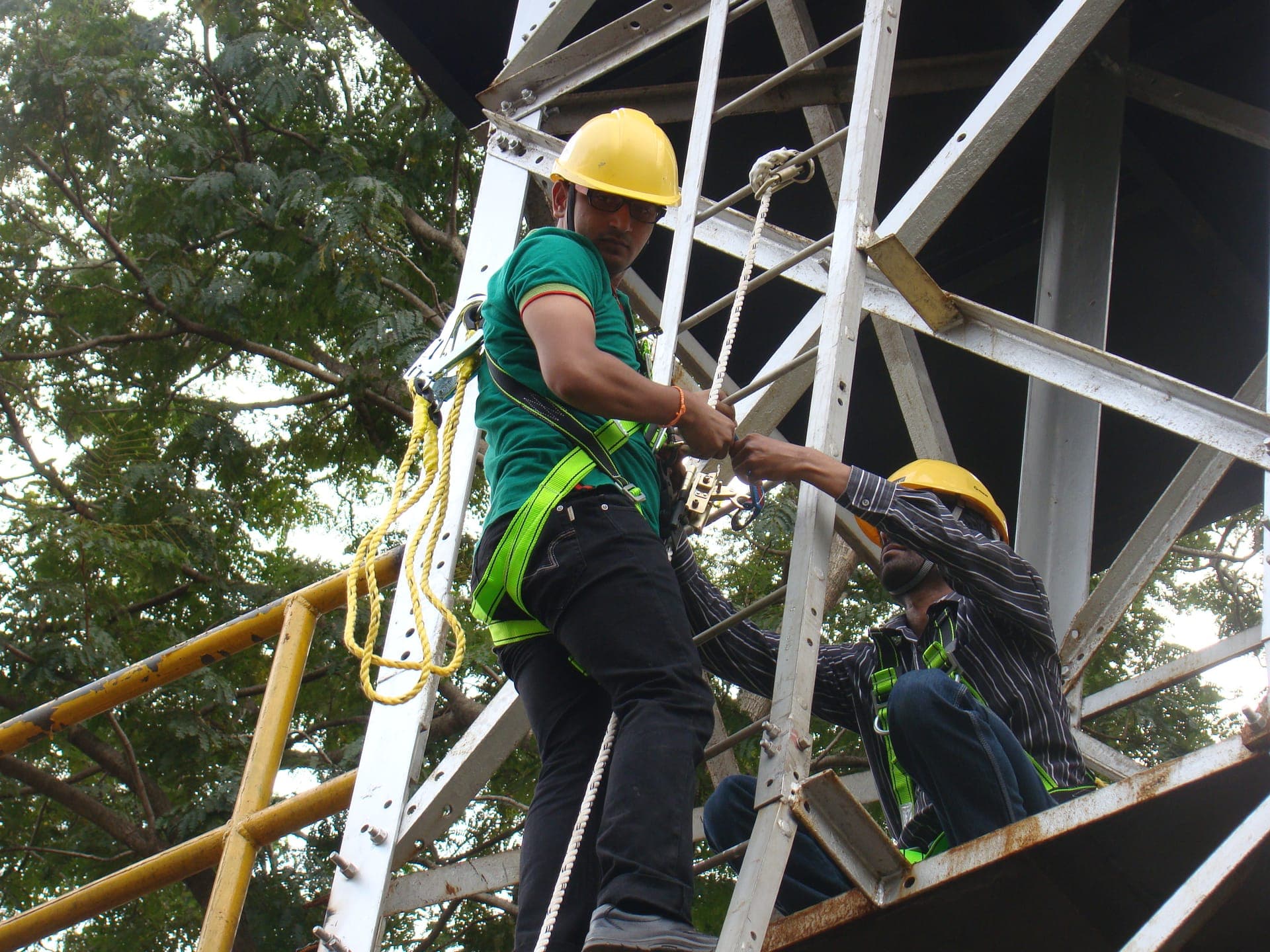 Trainee in a hard hat and full-body harness climbing a steel training structure during a work at height practical, with a second participant alongside